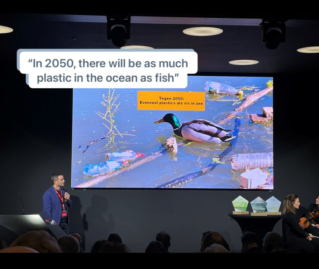 Boudewijn Decrop in front of a giant projector screen presenting a slide: Picture of duck in river full of plastic, title: "Tegen 2050: Evenveel plastics als vis in zee". Translation of slide title shown in image "In 2050, there will be as much plastic in the ocean as fish"